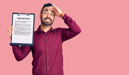 Young hispanic man holding clipboard with agreement document stressed and frustrated with hand on head, surprised and angry face