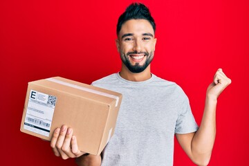 Young man with beard holding delivery package screaming proud, celebrating victory and success very excited with raised arm