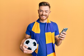 Young redhead man wearing hooligan scarf and using smartphone winking looking at the camera with...