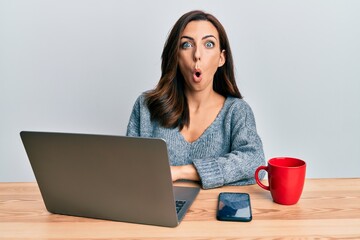 Young brunette woman working at the office using computer laptop scared and amazed with open mouth for surprise, disbelief face