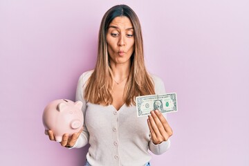 Brunette young woman holding one dollar banknote and piggy bank making fish face with mouth and squinting eyes, crazy and comical.