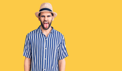 Young handsome man with beard wearing summer hat and striped shirt sticking tongue out happy with funny expression. emotion concept.
