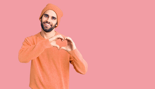 Young handsome man with beard wearing casual sweater and wool cap smiling in love showing heart symbol and shape with hands. romantic concept.