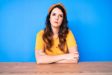 Young beautiful brunette woman sitting on the table over blue background skeptic and nervous,...