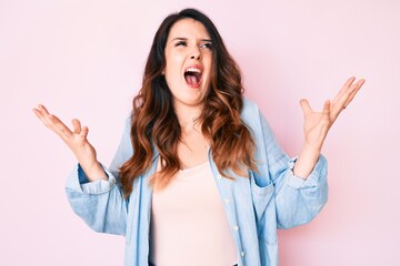 Young beautiful brunette woman wearing casual clothes over pink background crazy and mad shouting and yelling with aggressive expression and arms raised. frustration concept.