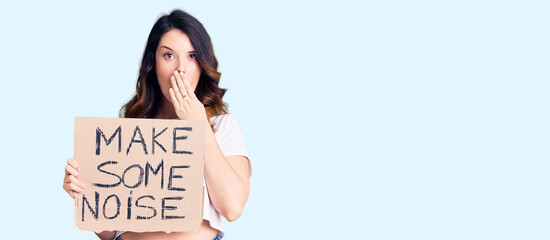 Beautiful young brunette woman holding make some noise banner covering mouth with hand, shocked and...