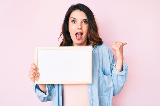Young beautiful brunette woman holding blank empty banner pointing thumb up to the side smiling happy with open mouth