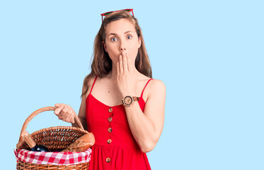 Young beautiful blonde woman holding picnic basket covering mouth with hand, shocked and afraid for...
