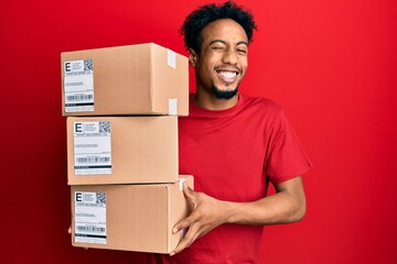 Young african american man with beard holding delivery packages winking looking at the camera with...