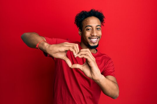 Young african american man with beard wearing casual red t shirt smiling in love doing heart symbol shape with hands. romantic concept.