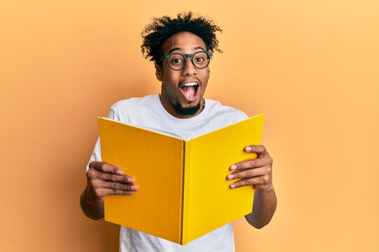Young african american man with beard reading a book wearing glasses celebrating crazy and amazed for success with open eyes screaming excited.