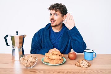 Young caucasian man with curly hair sitting on the table having breakfast smiling with hand over ear listening and hearing to rumor or gossip. deafness concept.