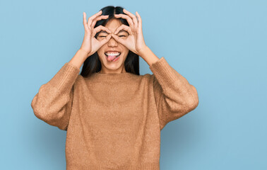 Beautiful young asian woman wearing casual winter sweater doing ok gesture like binoculars sticking tongue out, eyes looking through fingers. crazy expression.