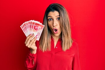 Beautiful brunette woman holding 20 israel shekels banknotes scared and amazed with open mouth for surprise, disbelief face