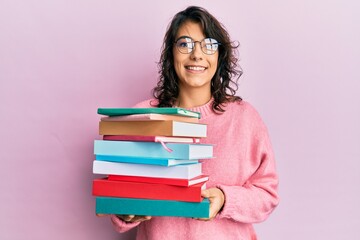 Young hispanic woman holding a pile of books looking positive and happy standing and smiling with a confident smile showing teeth