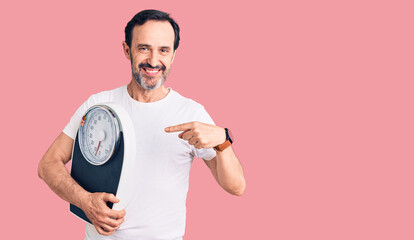 Middle age handsome man holding weighing machine pointing finger to one self smiling happy and proud