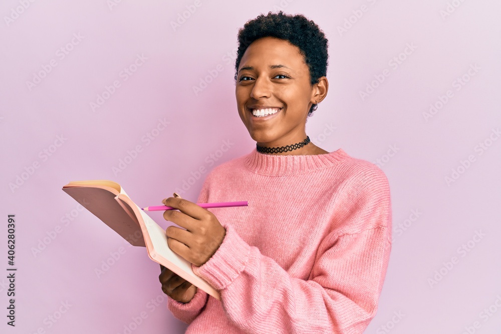 Wall mural Young african american girl reading and writing book smiling with a happy and cool smile on face. showing teeth.