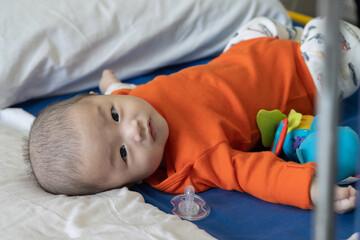 Illness asian baby boy lying down on sickbed and looking at camera, admitted in hospital and saline intravenous (IV) drip on hand.