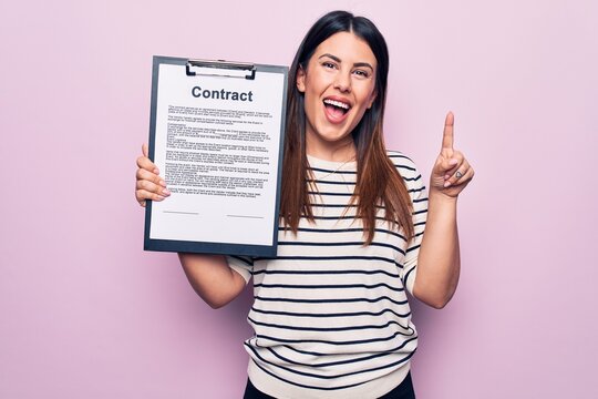 Young beautiful woman holding clipboard with contract document over pink background smiling with an idea or question pointing finger with happy face, number one