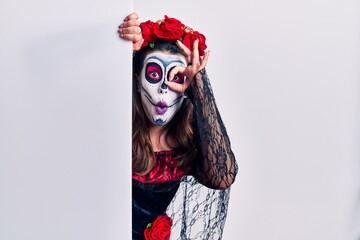 Young woman wearing day of the dead custome holding blank empty banner doing ok gesture shocked with surprised face, eye looking through fingers. unbelieving expression.