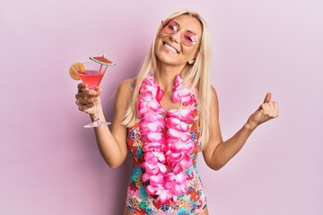 Young blonde woman wearing swimsuit and hawaiian lei drinking cocktail screaming proud, celebrating...