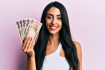 Beautiful hispanic woman holding japanese yen banknotes looking positive and happy standing and smiling with a confident smile showing teeth