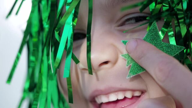 Adorable happy brown eyes child in green wig looking at the camera, while mother sticks green star on her cheek, celebrating saint patrick's day, white wall background. Extreme close up
