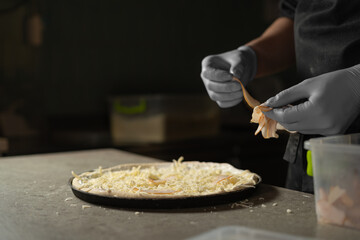 Chef's hands in gloves and a round pizza sprinkled with grated cheese. A baking dish on a table in production.