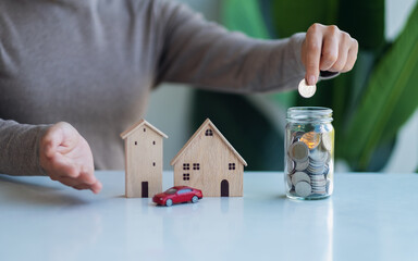 Closeup image of a woman collecting and putting coins in a glass jar with wooden house models and...