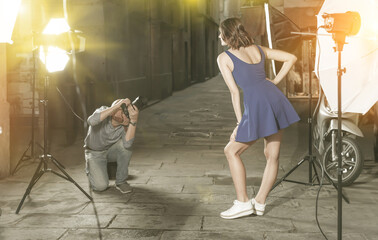 Photographer using professional camera and light equipment for taking pictures of cheerful woman in blue dress on town street