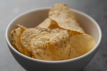 White bowl with organic potato chips sprinkled with fresh ground black pepper on concrete background