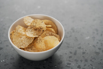 White bowl with organic potato chips sprinkled with fresh ground black pepper on concrete background with copy space