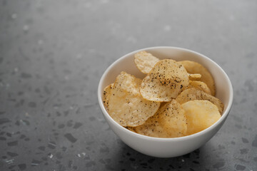 White bowl with organic potato chips sprinkled with fresh ground black pepper on concrete background with copy space