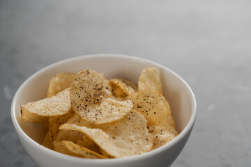 White bowl with organic potato chips sprinkled with fresh ground black pepper on concrete background with copy space