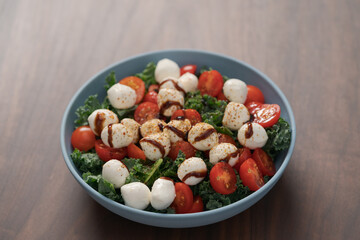 Blue bowl of salad with mozzarella, cherry tomatoes and kale, seasoned with balsamic sauce on walnut wood table