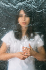 Fototapeta na wymiar Plastic pollution. Defocused silhouette. Garbage collection. Zero waste. Volunteer woman holding used bottles behind wrinkled texture polyethylene film isolated on dark background out of focus.