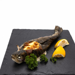 Whole baked and stuffed trout. Decoration - sprigs of curly parsley, lemon wedges. Lies on a black stone serving tile. White background.