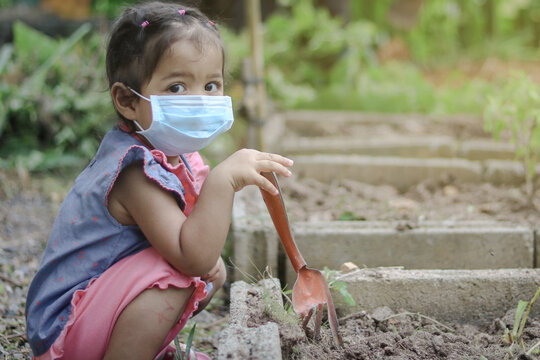 Asian Little Girl Is Planting The Plant In The Garden And Wearing Surgical Mask Outside The House, Concept Of Dust PM 2.5, Covid-19, Coronavirus Protection. 