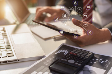 Designer using tablet with laptop and calculator on desk in office with virtual interface graphic icons network diagram.