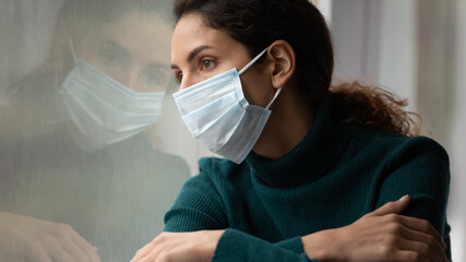 Upset young Caucasian woman in medical facial mask look in window feel lonely distressed sick with covid-19. Unhappy female in facemask suffer from coronavirus. Quarantine, corona pandemic concept. - obrazy, fototapety, plakaty