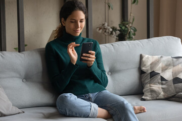 Obraz premium Millennial Caucasian woman relax on sofa in living room using modern cellphone gadget. Young female rest on couch at home look at smartphone screen text message online. Technology concept.