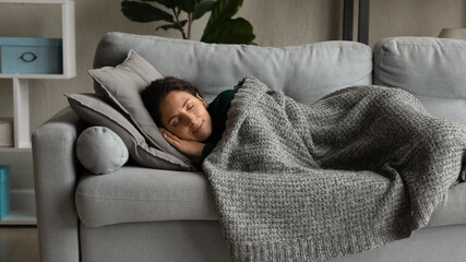 Happy millennial Caucasian woman lying under blanket on cozy couch at home sleeping or dreaming. Smiling calm young female relax rest on sofa in living room, relieve negative emotions daydreaming. - obrazy, fototapety, plakaty