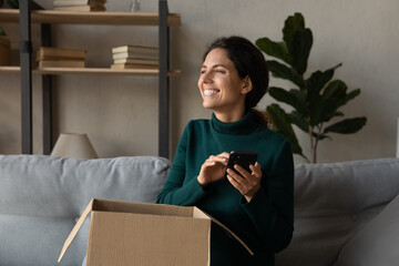 Smiling millennial Caucasian woman use cell track order shopping online unpacking cardboard box....