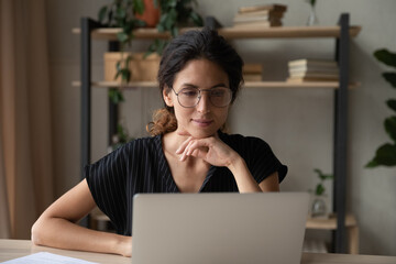 Happy millennial Caucasian woman in spectacles look at laptop screen work online on gadget from home office. Smiling young female use computer browse internet on device. Technology concept.