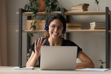 Smiling Caucasian woman in headphones wave greet to camera talking on video call on laptop. Happy young female in glasses and earphones have webcam digital conference or meeting on computer.