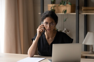 Focused serious young Caucasian businesswoman look at laptop screen talk on smartphone with customer. Millennial woman work on computer consult client online on cellphone from home office.