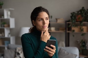 Pensive young Caucasian woman use smartphone look in distance thinking pondering. Thoughtful millennial female hold cellphone texting messaging make decision or plan. Technology concept.