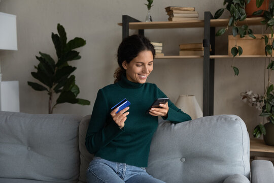 Happy millennial Caucasian woman sit on sofa at home shopping online on smartphone with credit card. Smiling young female relax on couch buy on internet use secure payment system.