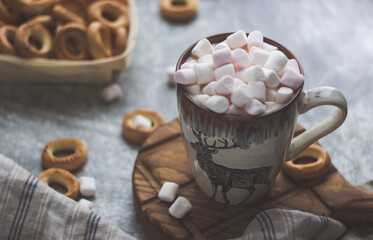 close-up of a mug of cocoa with pink marshmallows on a brown wooden stand, round bagels nearby; shot from above,gray toning