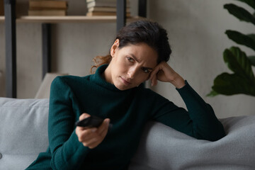 Obraz premium Unhappy young Caucasian woman sit rest on sofa at home bored at home watching television. Upset millennial female feel lazy dissatisfied choose TV program switch channel with remote controller.